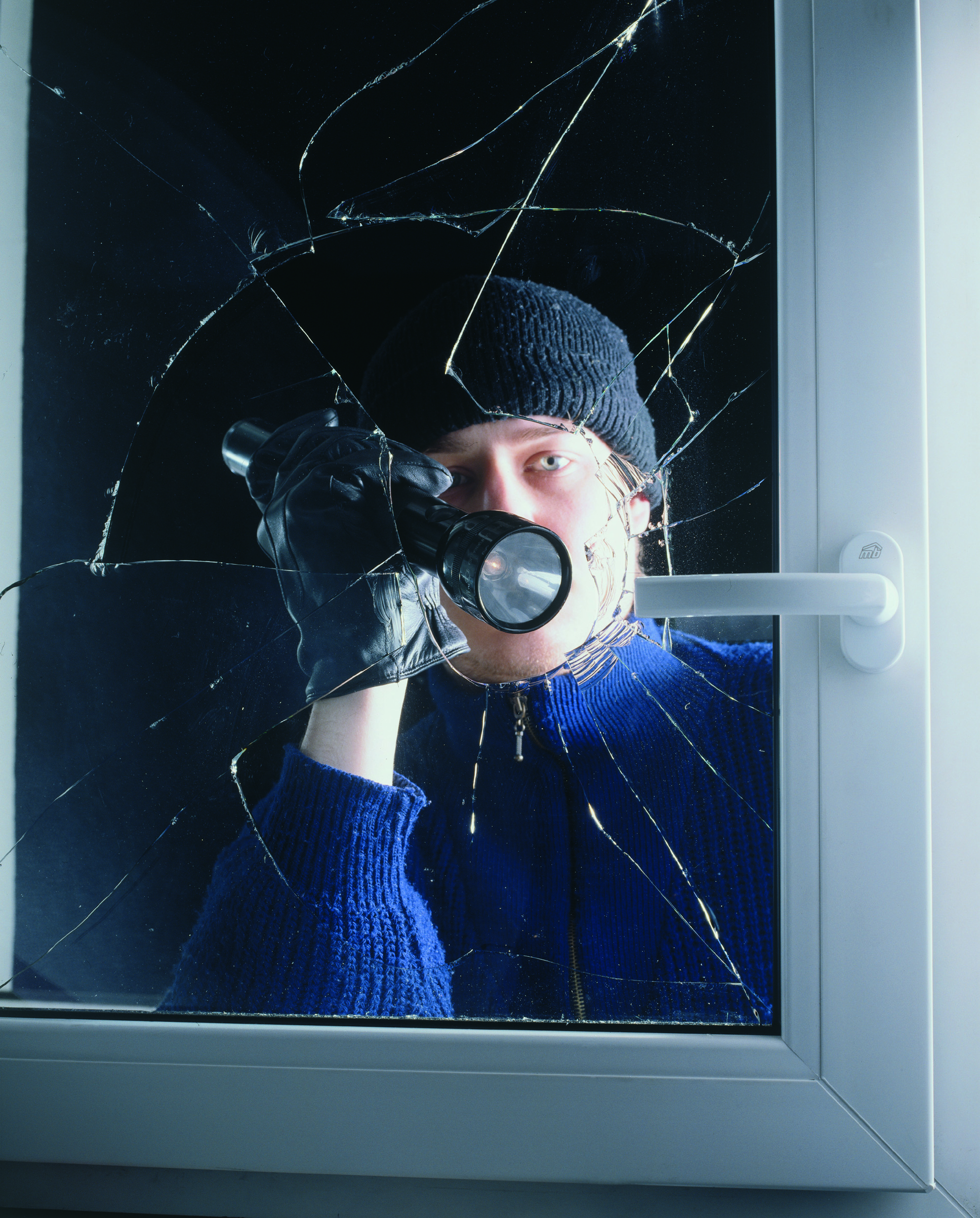 Foto: Initiative für aktiven Einbruchschutz „Nicht bei mir!" Einbrecher mit Taschenlampe leuchtet durch zerbrochenes Fenster